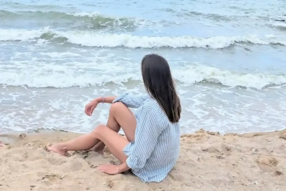 A woman sits by the sea and looks towards the waves.